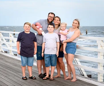 Family of six smiling on a seaside boardwalk, with an ocean view in the background.