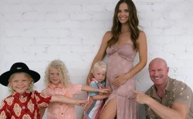 Family photo with a pregnant woman, three smiling children, and a man in front of a white brick wall.