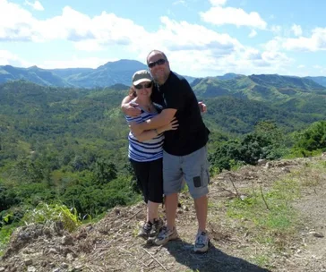 A couple hugging on a hilltop with a scenic view of green mountains and a bright blue sky in the background.