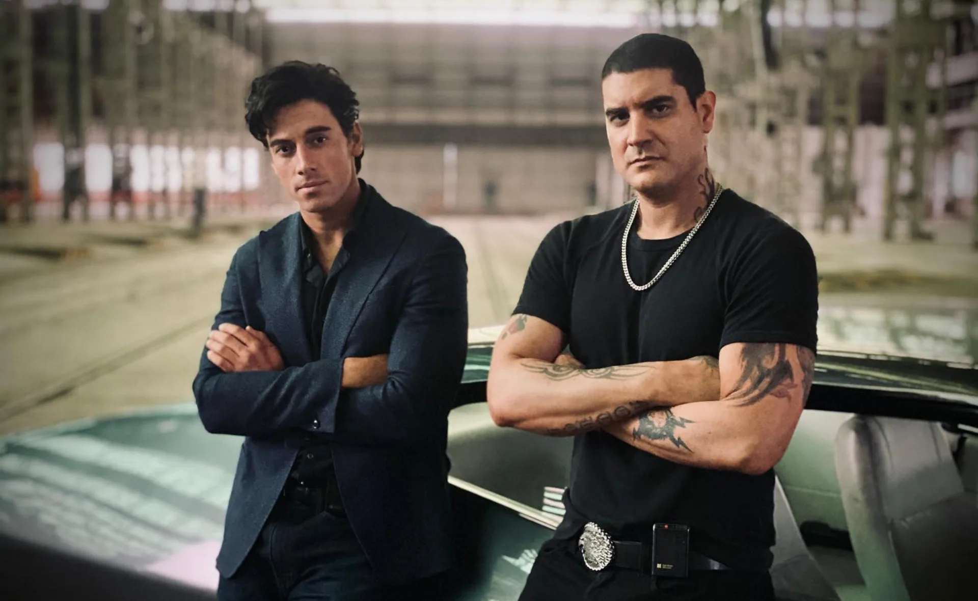 Two men stand confidently by a car in an industrial setting; one wears a suit, the other a black t-shirt with tattoos.
