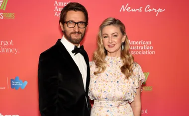 A couple posing at an event in front of a red backdrop featuring various logos.