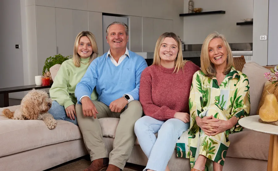 Family of four smiling on a couch with a dog in a cozy living room setting.