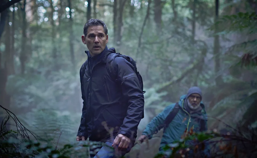 Two men hike through a dense forest, wearing backpacks and outdoor gear, looking alert and cautious.