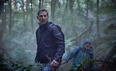 Two men hike through a dense forest, wearing backpacks and outdoor gear, looking alert and cautious.