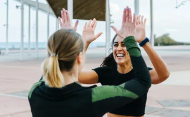 Two women high-five each other outdoors, smiling broadly in athletic attire.