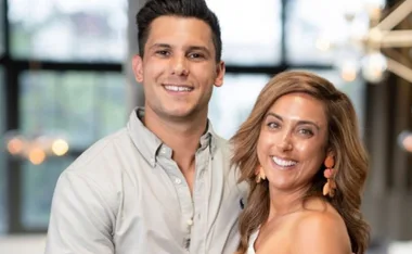 A smiling couple stands together indoors with blurred lights in the background.