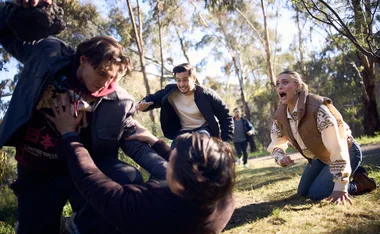 A chaotic scene with people fighting and a woman kneeling, appearing distressed, in a forest setting.