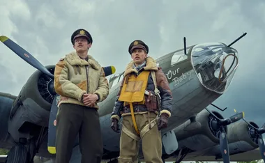 Two men in WWII pilot uniforms stand confidently in front of a vintage bomber plane labeled "Our Baby."