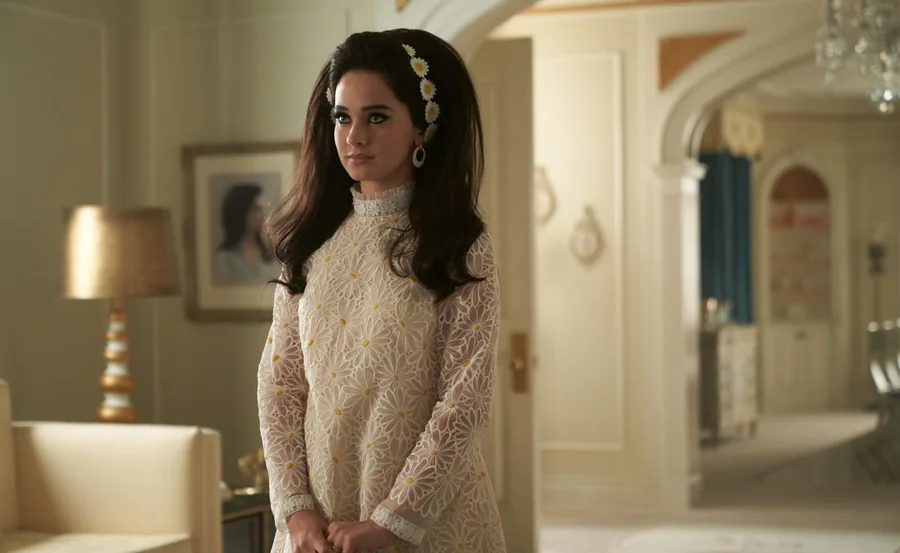 Woman in a 1960s-style floral dress with bouffant hair and daisy headband, standing in a vintage room.