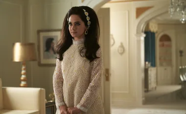 Woman in a 1960s-style floral dress with bouffant hair and daisy headband, standing in a vintage room.