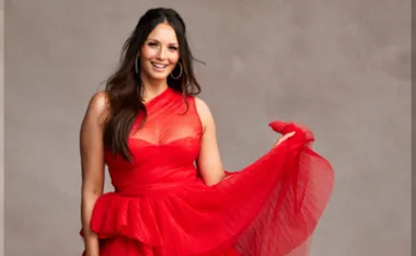 A woman in a flowing red dress poses against a neutral background, smiling with long brown hair and hoop earrings.