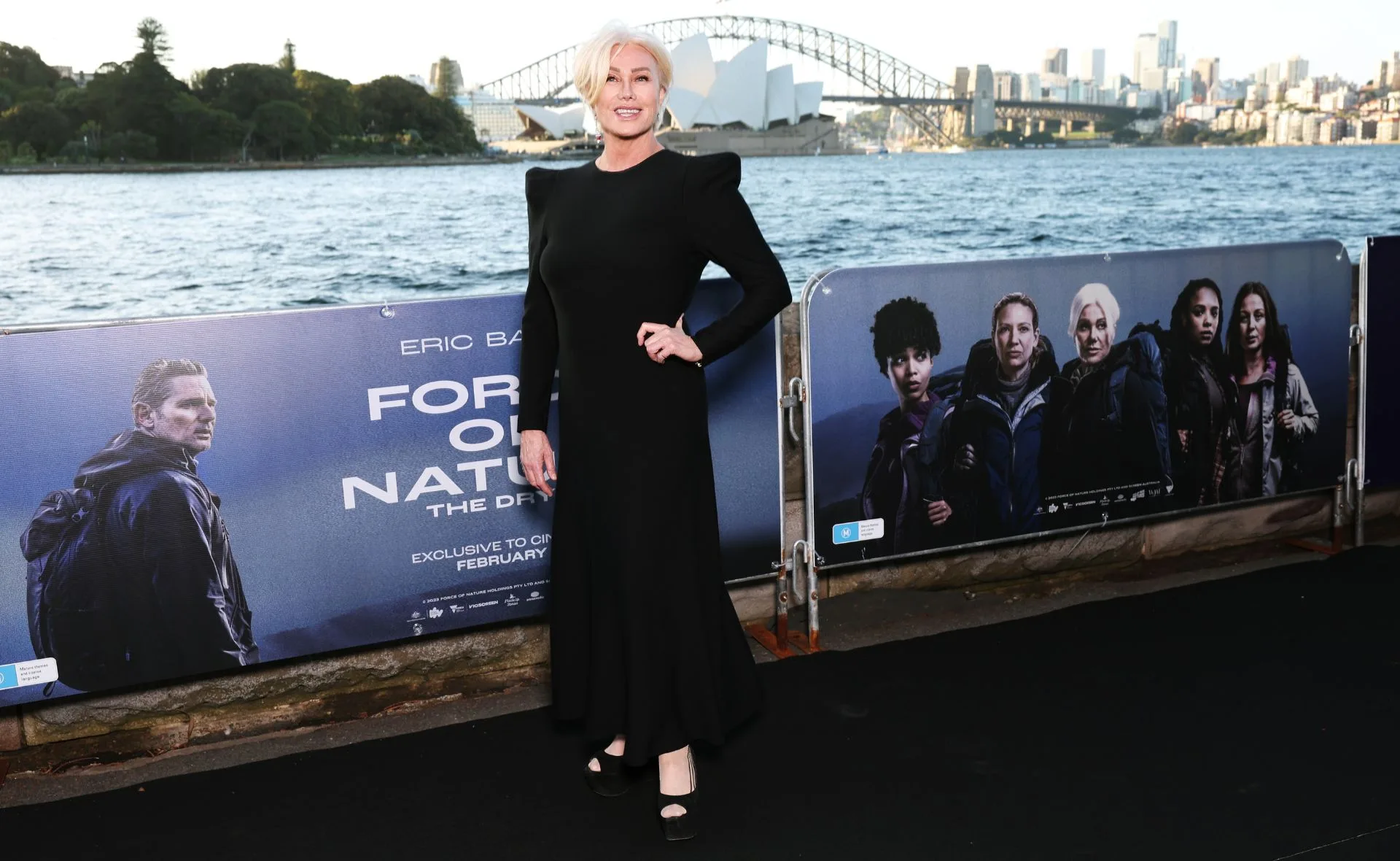 A woman in a black dress stands by a promotional poster for "Force of Nature" near Sydney Opera House.
