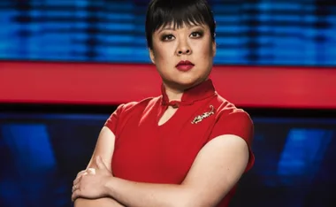 A person in a red dress with short black hair stands confidently against a blurred background.