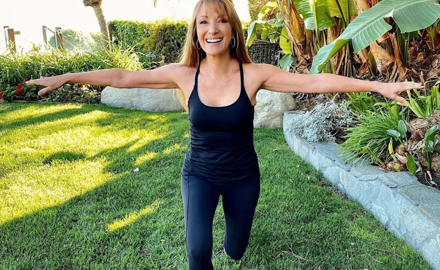 A woman in black workout attire smiles brightly while exercising in a lush green garden.