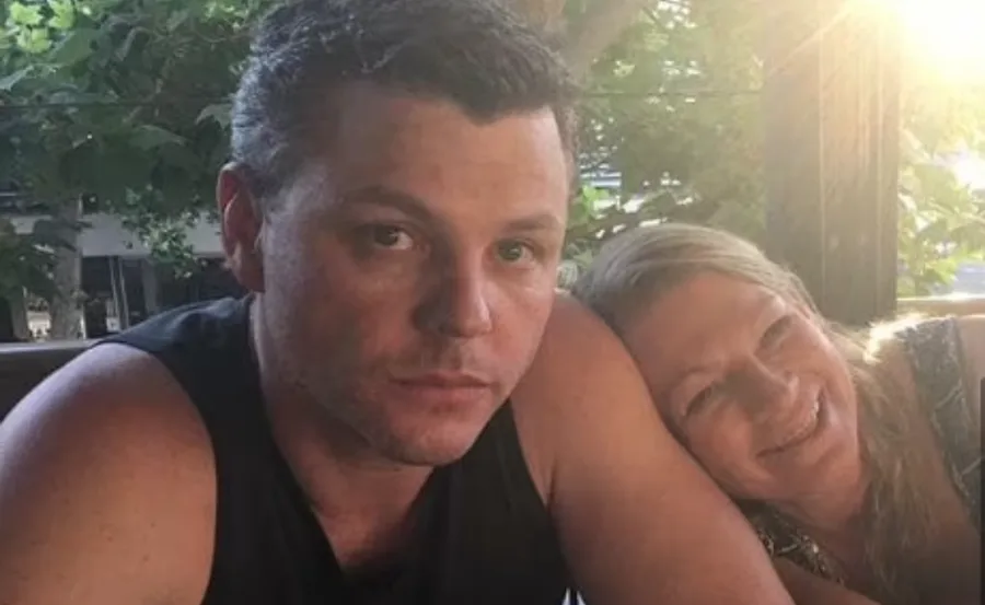 Two people sitting at a table outdoors; the man looks at the camera while the woman rests her head on his shoulder, smiling.