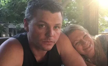 Two people sitting at a table outdoors; the man looks at the camera while the woman rests her head on his shoulder, smiling.