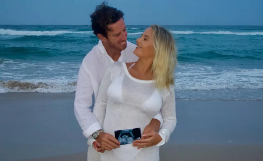 Couple in white outfits on a beach holding an ultrasound photo, smiling at each other.