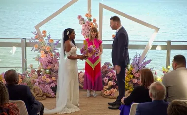 A bride and groom stand at the altar with a celebrant, surrounded by colorful flowers, overlooking the ocean.