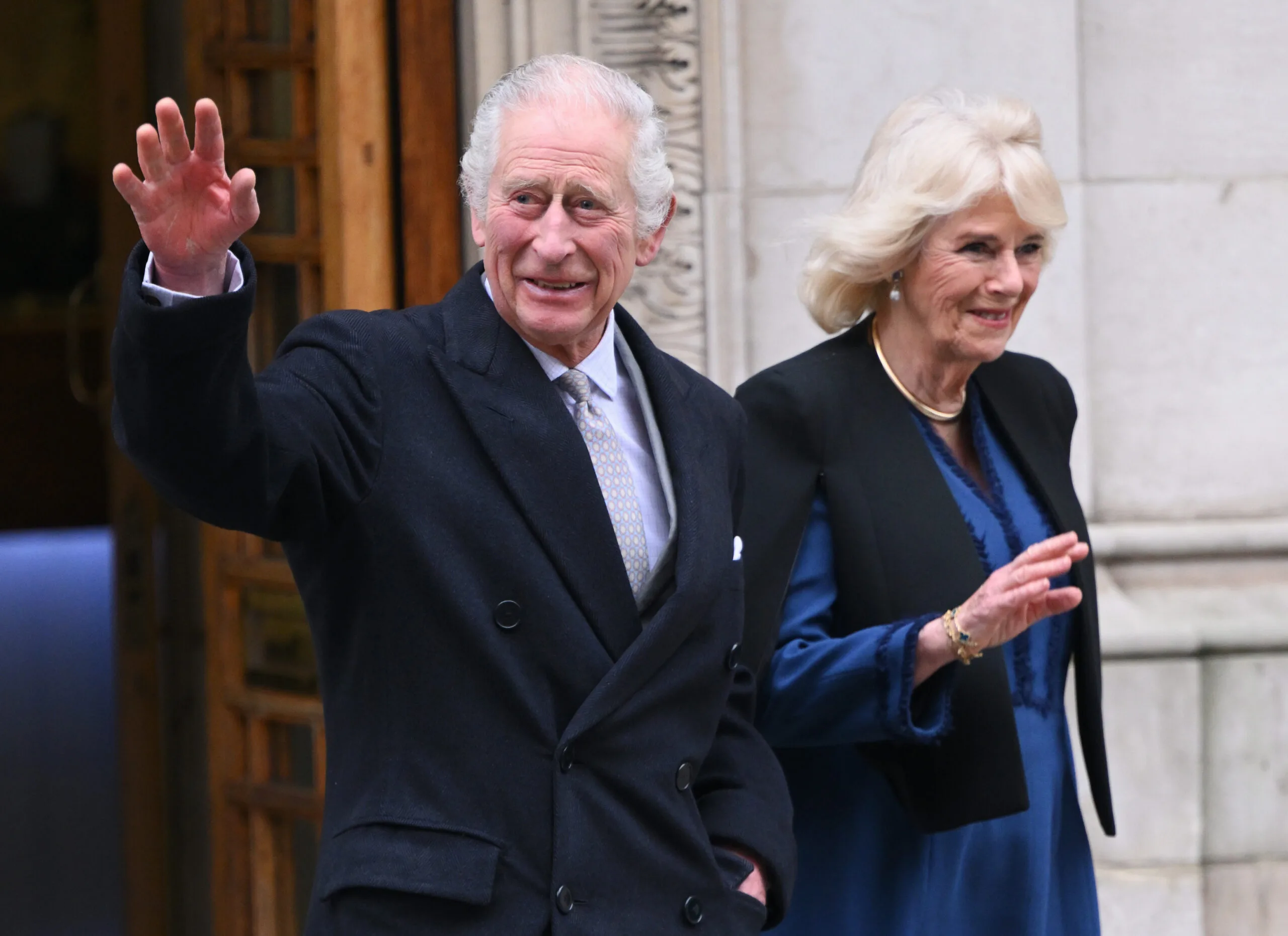 Man and woman waving outside a building, both smiling warmly and dressed in formal attire.