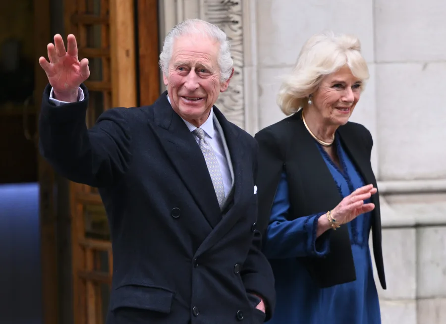 Man and woman waving outside a building, both smiling warmly and dressed in formal attire.
