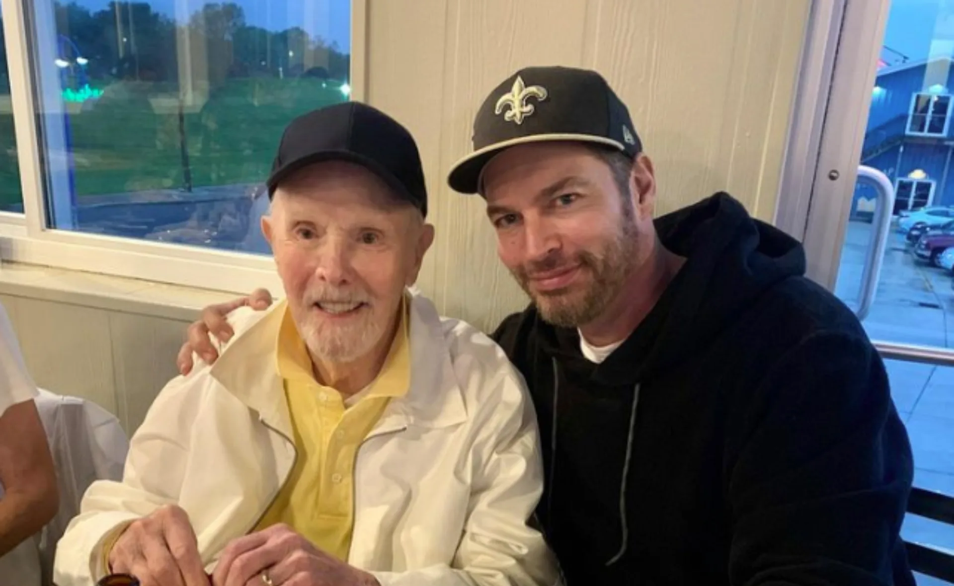 Two men smiling indoors, both wearing baseball caps; one in a white jacket, the other in a black hoodie.