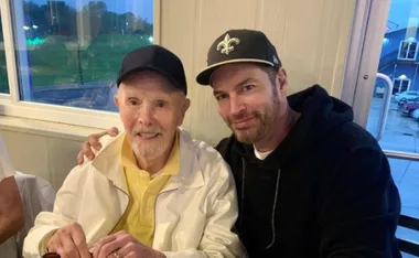 Two men smiling indoors, both wearing baseball caps; one in a white jacket, the other in a black hoodie.