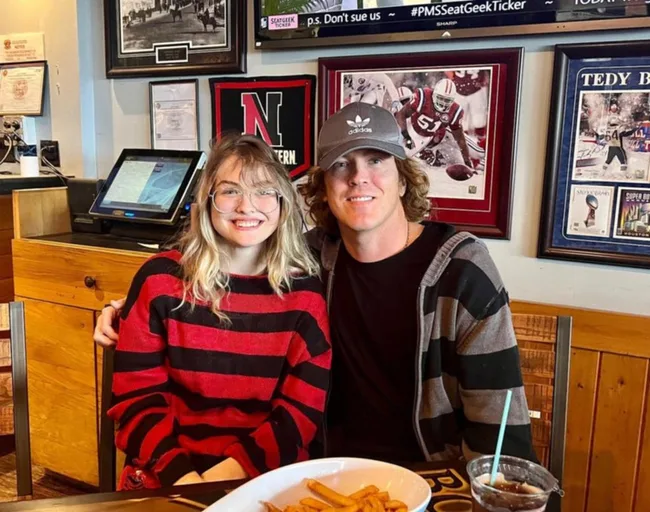 A young woman in a red and black striped sweater sits with a man in a cap at a restaurant, smiling at the camera.