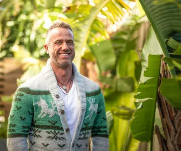 Man smiling in a patterned cardigan, standing outdoors with lush green plants in the background.