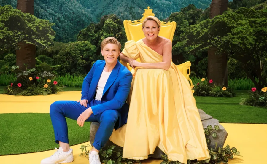 Two people in a colorful set, one sitting in a yellow gown on a throne, the other in a blue suit seated on a rock.