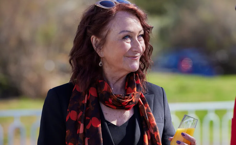 Elderly woman with sunglasses on head and colorful scarf smiles holding a glass of orange juice outdoors.