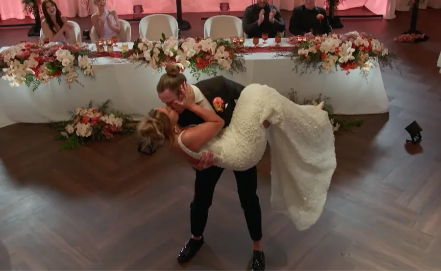 Couple in wedding attire, partner dips and kisses bride on dance floor, guests applauding in background.