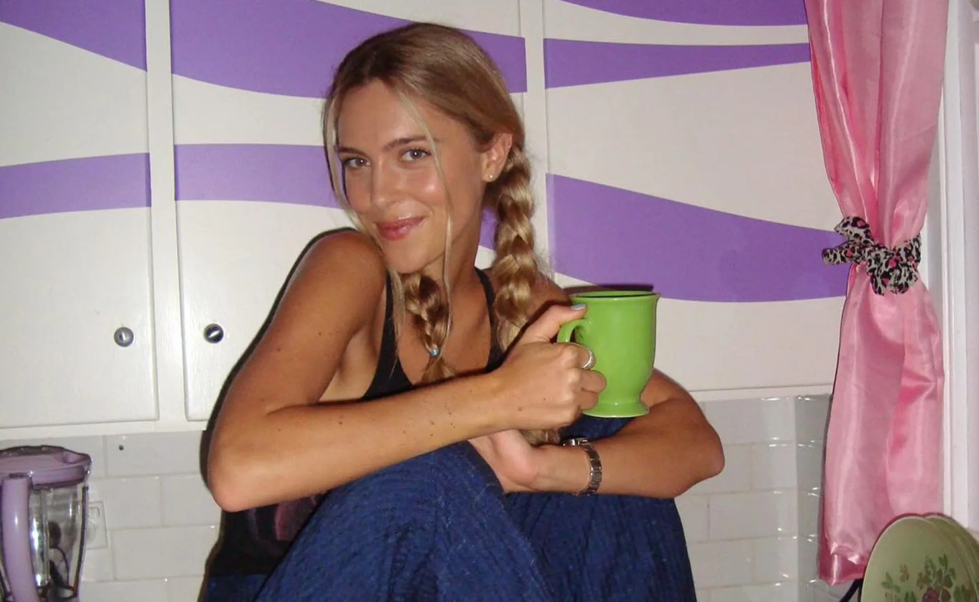 Young woman with braided hair holding a green mug in a kitchen with purple and white cabinets and pink curtains.