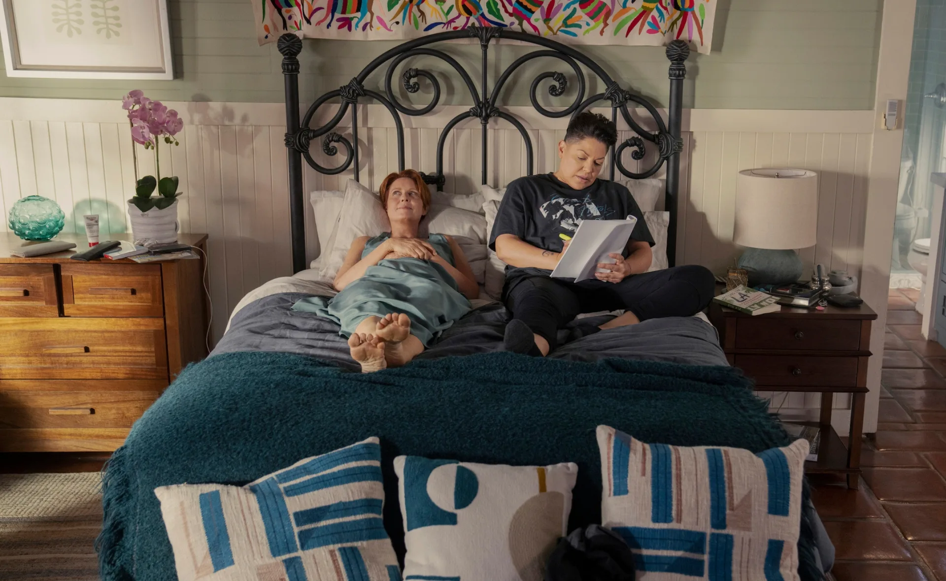 Two people relaxing on a bed, one reading, in a cozy, colorful bedroom setting.