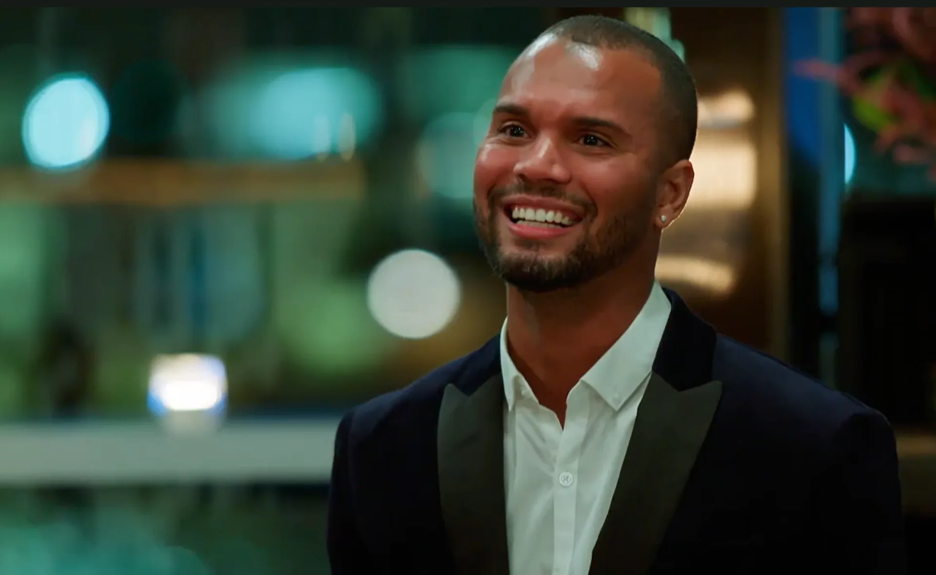 A person in a suit smiles warmly, standing in a softly lit indoor setting.