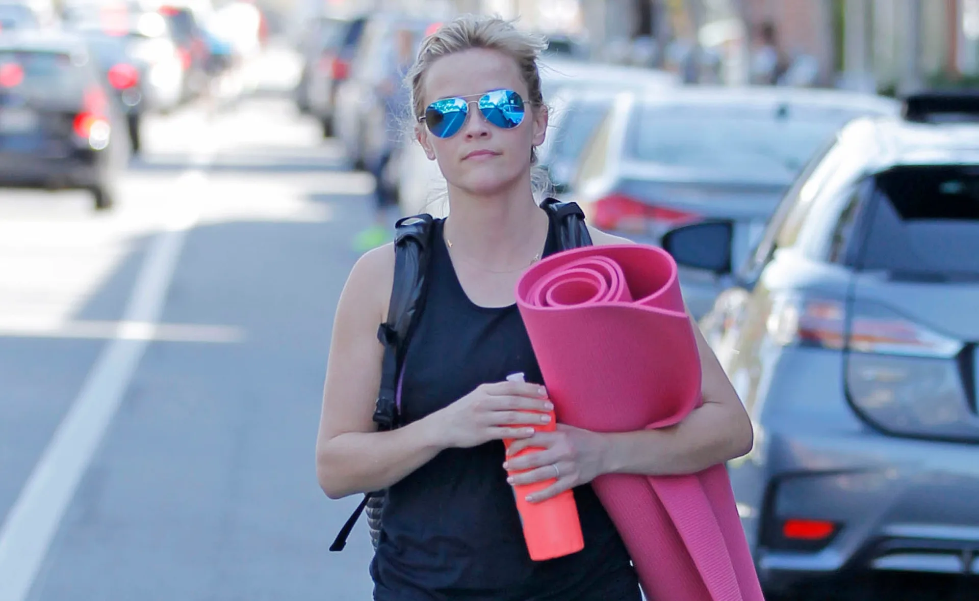 Person wearing sunglasses carrying a pink yoga mat and an orange water bottle on a city street.