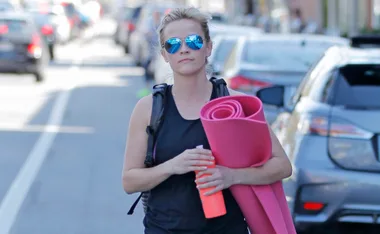 Person wearing sunglasses carrying a pink yoga mat and an orange water bottle on a city street.