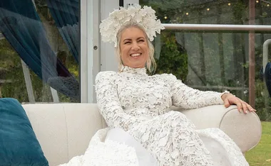 A woman in a white lace dress and floral headpiece smiles while sitting on a couch outdoors.