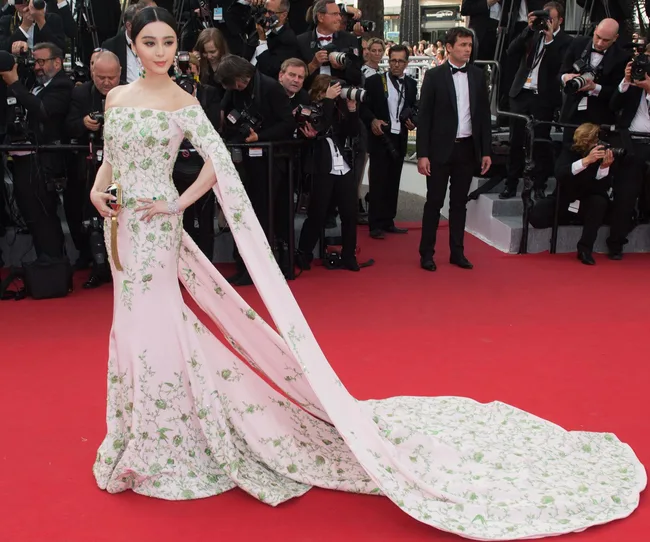 A woman in a floral gown with a long train poses on a red carpet surrounded by photographers.