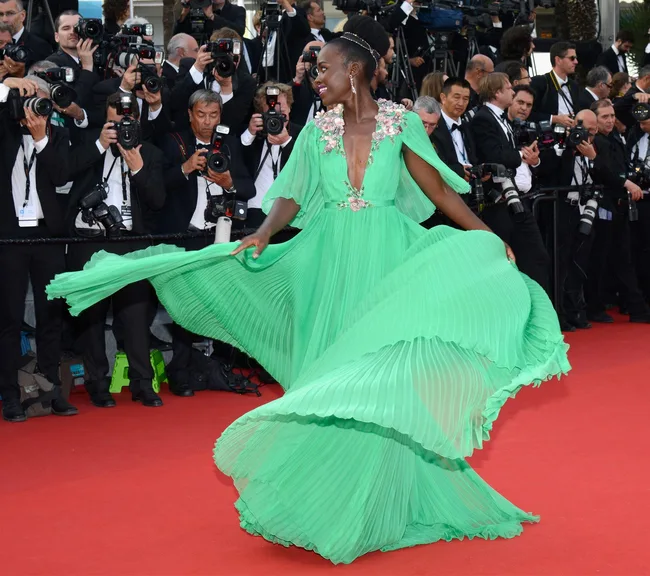 Dazzling person in a flowing green dress twirls on the red carpet amidst photographers at a glamorous event.
