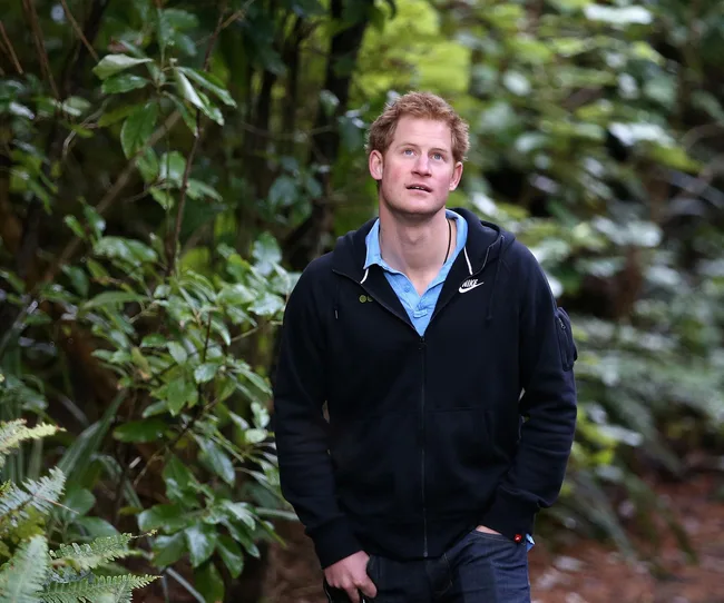 Young man with red hair in a black hoodie walking through a forest, looking upwards thoughtfully.