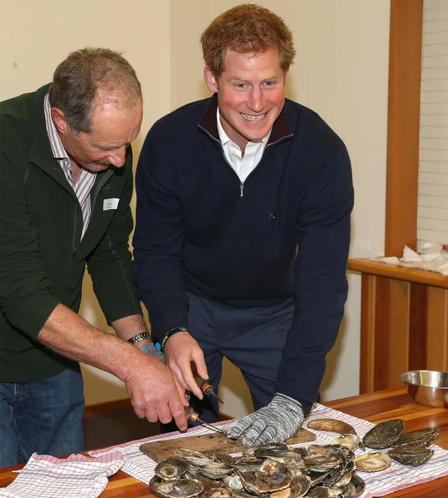 Two people are smiling and shucking oysters at a table covered with a towel and a bowl of shells.