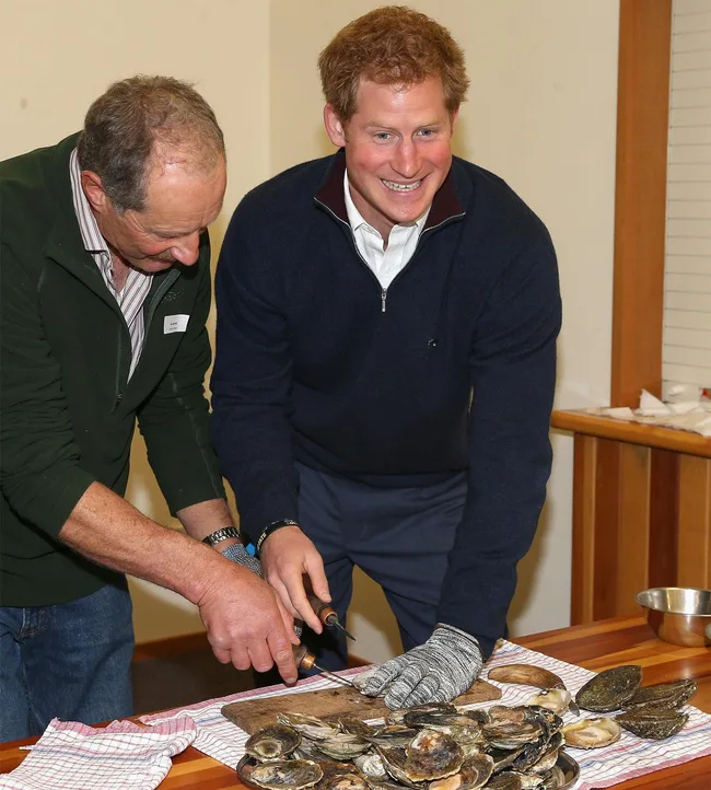 Two people are smiling and shucking oysters at a table covered with a towel and a bowl of shells.