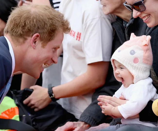 "Man in suit smiles at a baby wearing a cute hat, surrounded by a crowd."