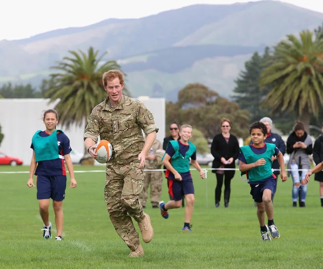 Man in military uniform playing rugby with children on a grassy field, trees and hills in the background.