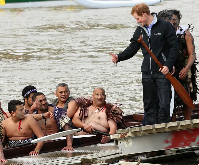 Man standing on dock holding a paddle, giving a thumbs-up to a group in a traditional canoe on the water.