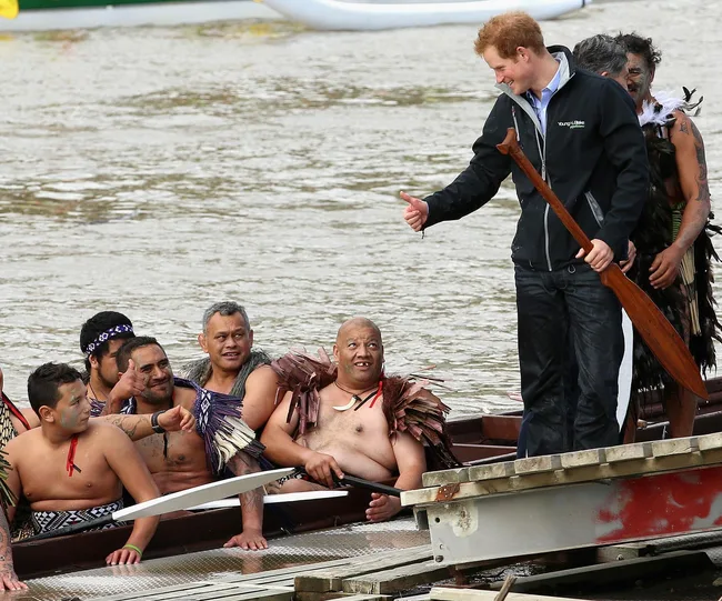 Man standing on dock holding a paddle, giving a thumbs-up to a group in a traditional canoe on the water.