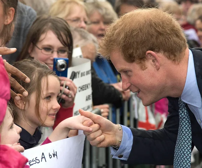 A man in a suit smiling and greeting a young girl in a crowd, with onlookers holding cameras and signs in the background.