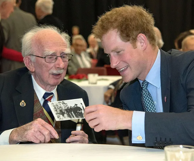 An elderly man and a younger man smile while looking at a black-and-white photo at a table in a social gathering.
