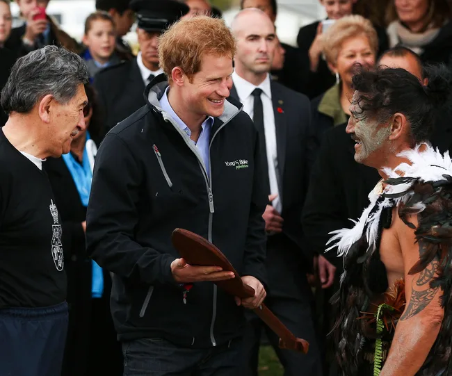Three men smiling; one in traditional attire with feathers, others in modern clothing; people in background.