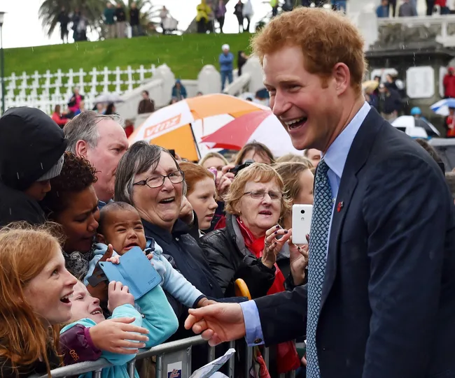 A person in a suit greets a crowd of smiling people, including children, while it rains. Some people hold umbrellas.